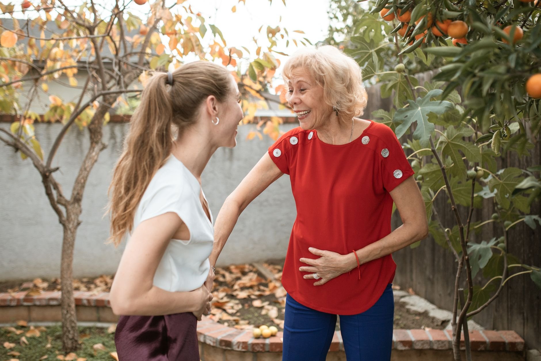 Two women laughing and talking together in a garden, enjoying a sunny day.